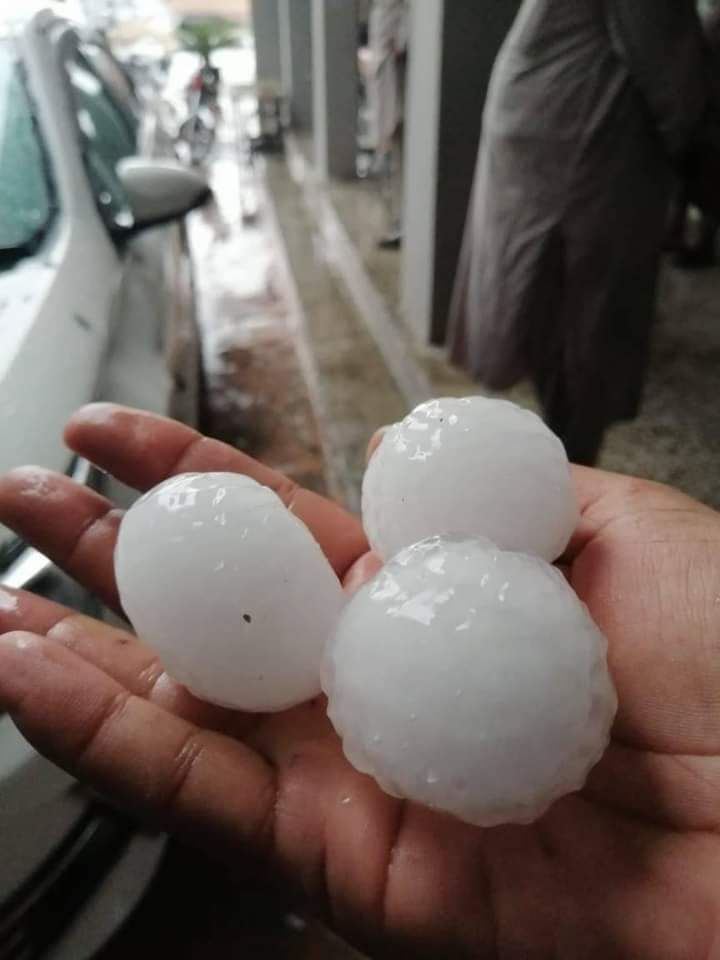 A hand holding three large hailstones beside parked vehicles.